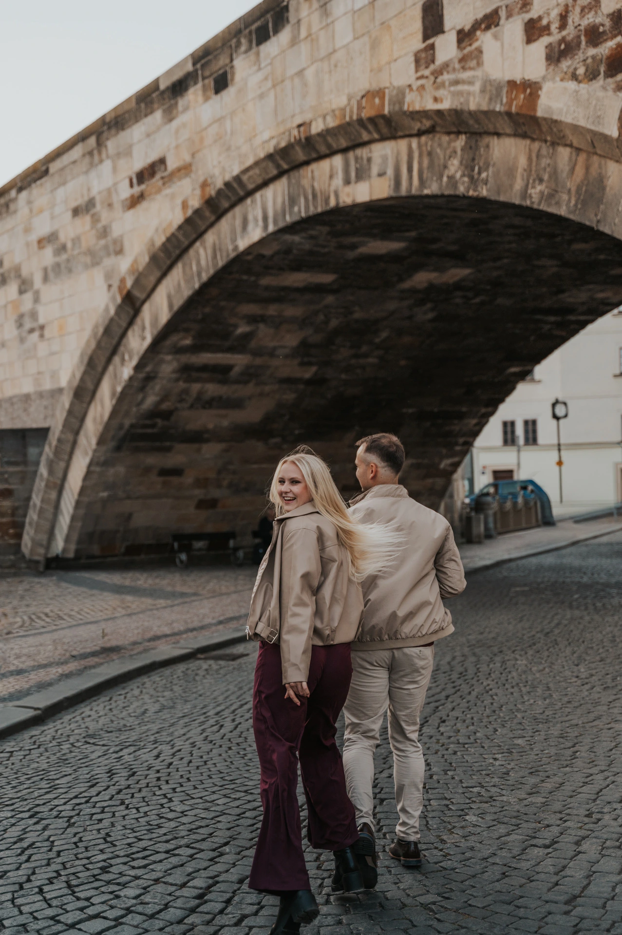 Couple looking back while walking beneath Charles Bridge during a couple photoshoot in Prague.