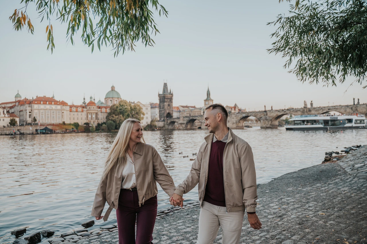 Prague couple photoshoot: couple smiling as they hold hands beside the river. Couple smiling as they hold hands beside the river during a couple photoshoot in Prague.