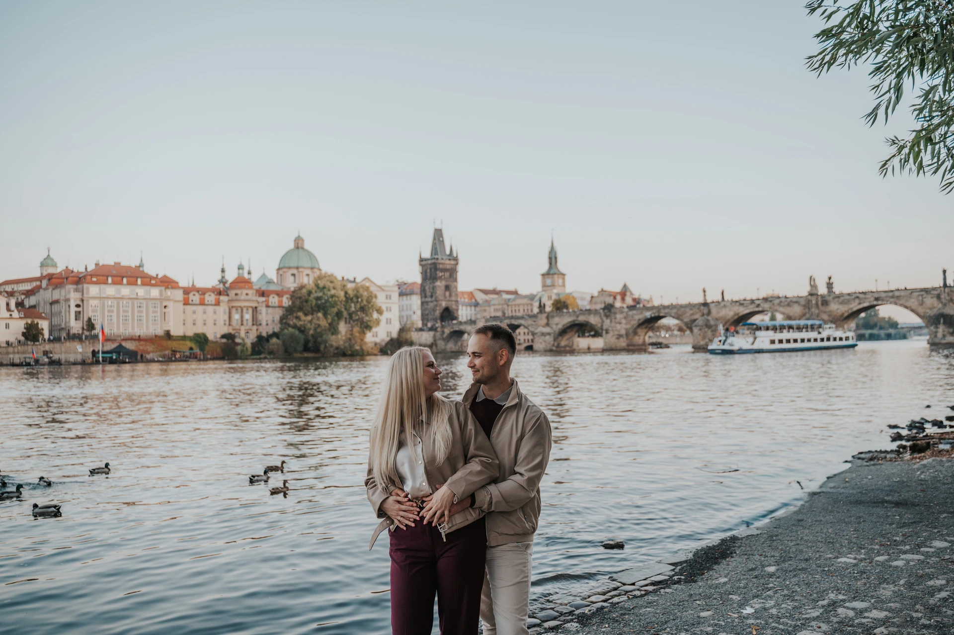 Wide portrait of the couple holding each other on the riverbank during a couple photoshoot in Prague.