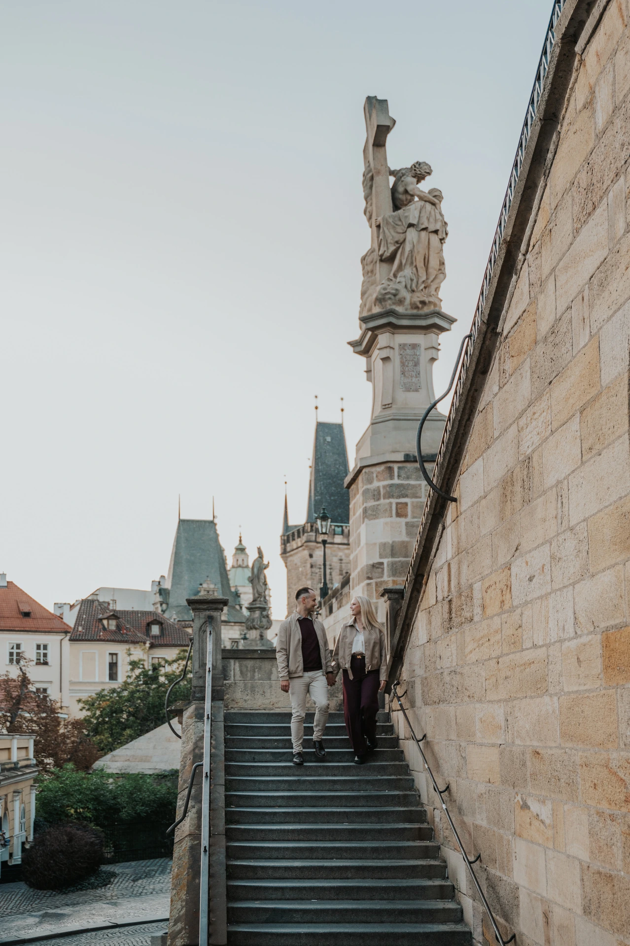 Wide view of the couple descending the bridge-side staircase during a couple photoshoot in Prague.