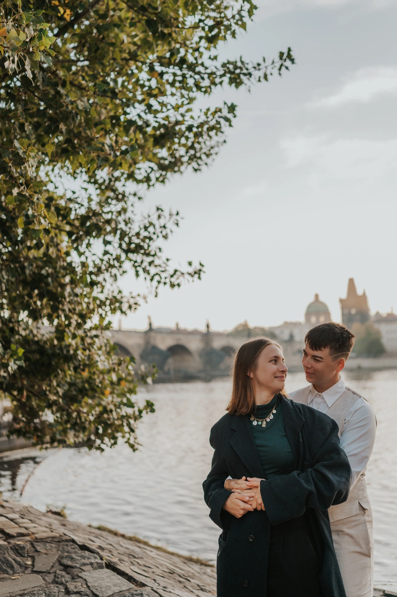 Couple embracing beneath the riverside trees during a couple photoshoot in Prague.