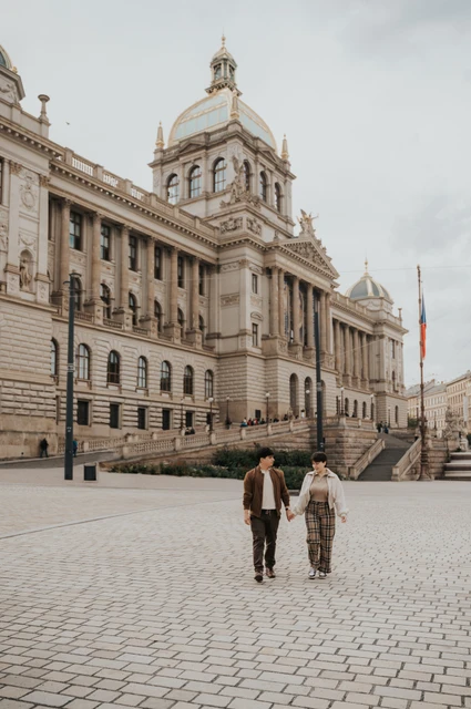 Prague couple photoshoot: wide view of the couple walking with the National Museum behind them. Wide view of the couple walking with the National Museum behind them during a couple photoshoot in Prague.