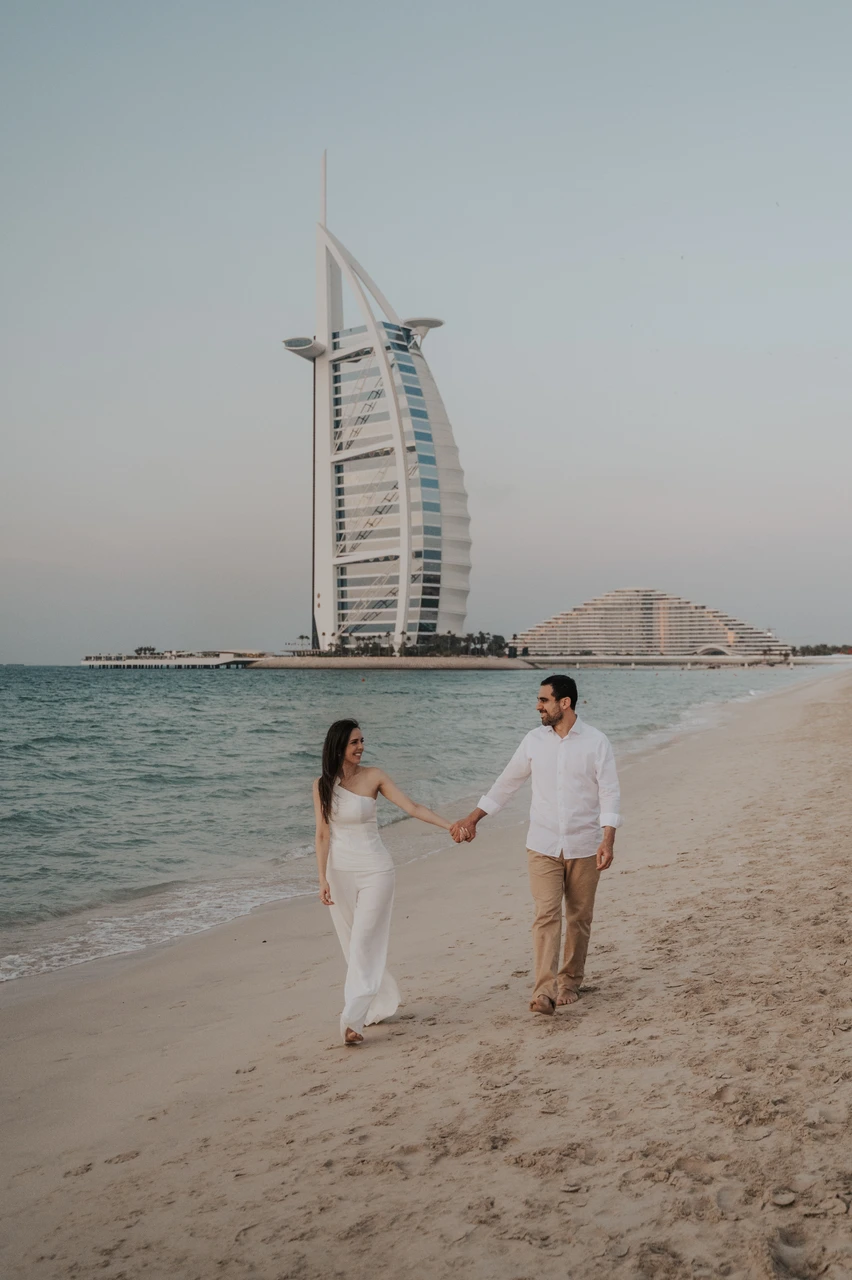 Dubai Proposal Costs - Burj Al Arab couple walk A couple walks hand in hand on Dubai beach with Burj Al Arab behind them after their proposal, captured in a proposal photoshoot.