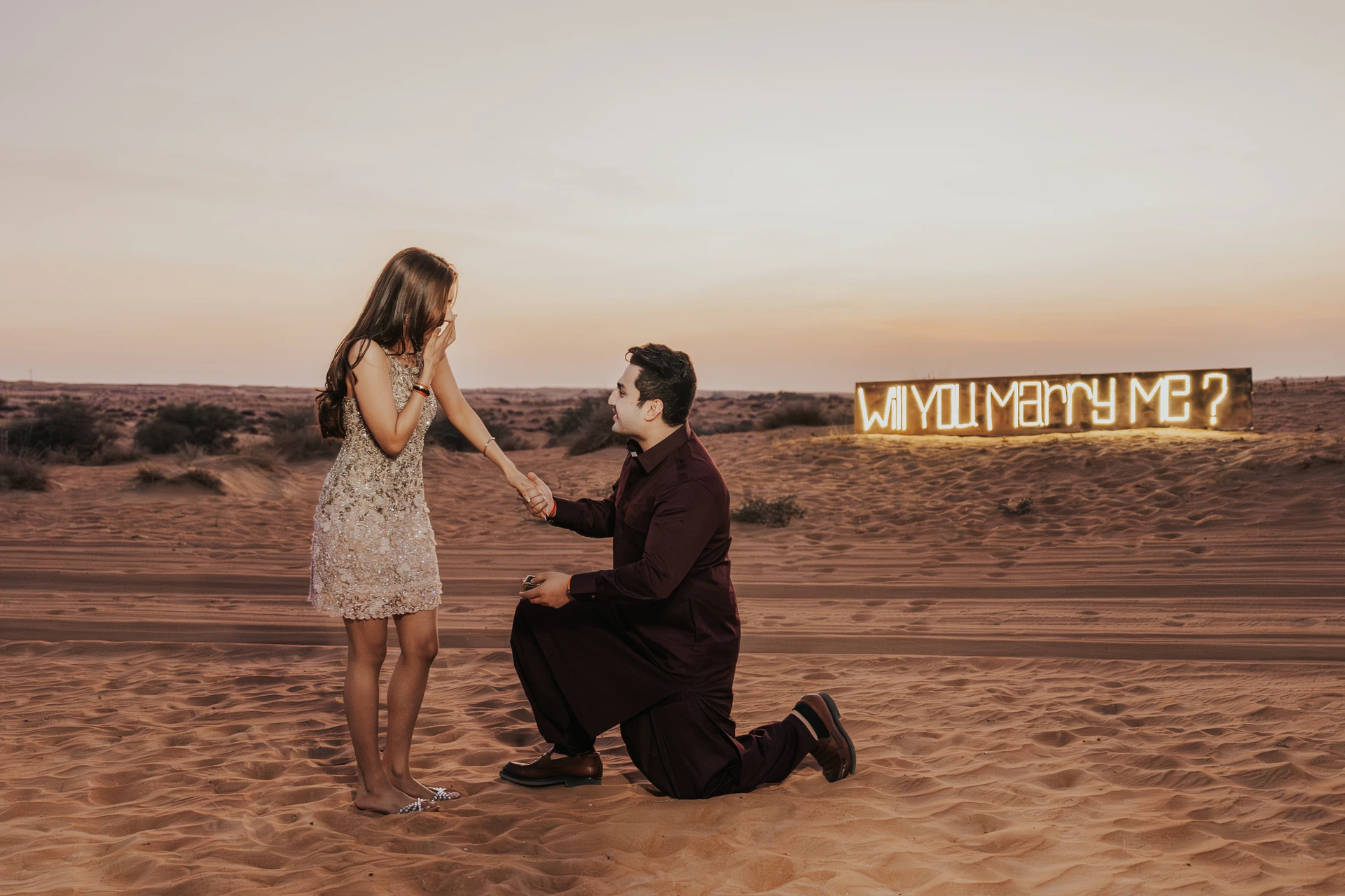 A man kneels on desert sand in Dubai with a Marry Me sign behind him, proposing during a styled evening proposal photoshoot.