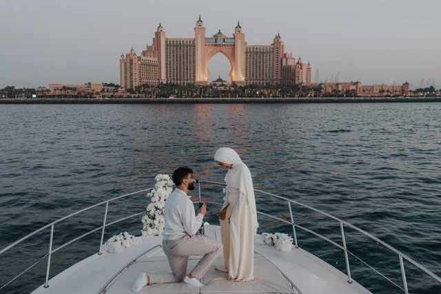 A man kneels on the bow of a yacht to propose in Dubai with Atlantis The Palm visible behind them during a proposal photoshoot.