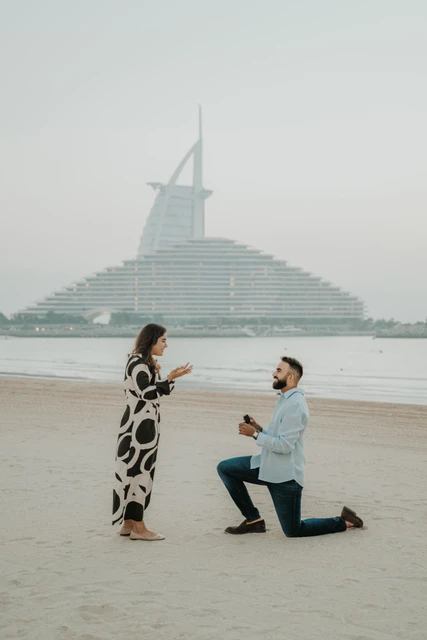 A man kneels with a ring box on a Dubai beach while his partner smiles, with Burj Al Arab behind them.