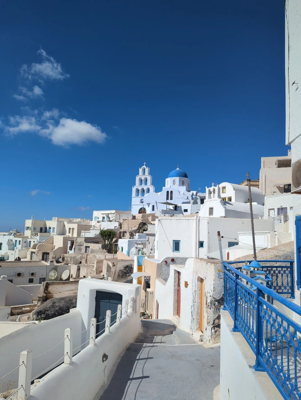 White Pyrgos lane descending past blue railings, with a blue-domed church and layered village rooftops ahead
