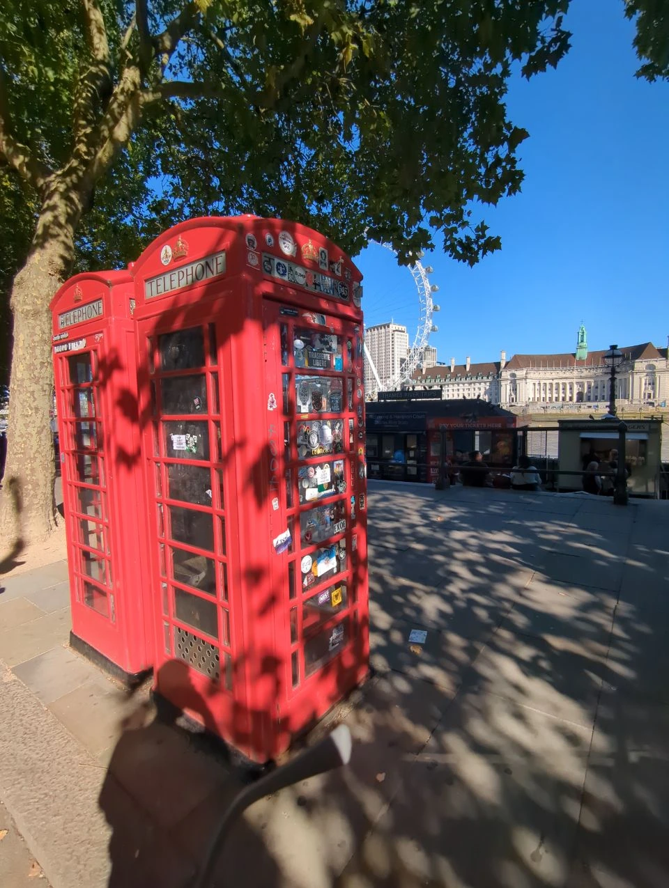 Two red telephone boxes with stickers beneath a leafy tree on Victoria Embankment, with part of the London Eye behind