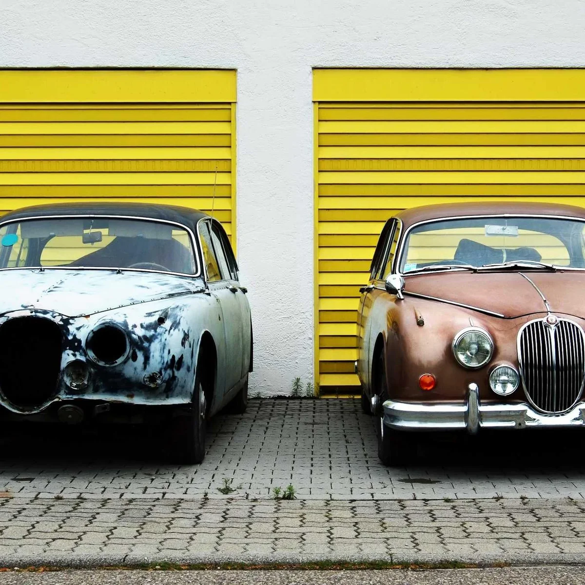 Two vintage cars, one rusted and one restored, parked in front of yellow garage doors