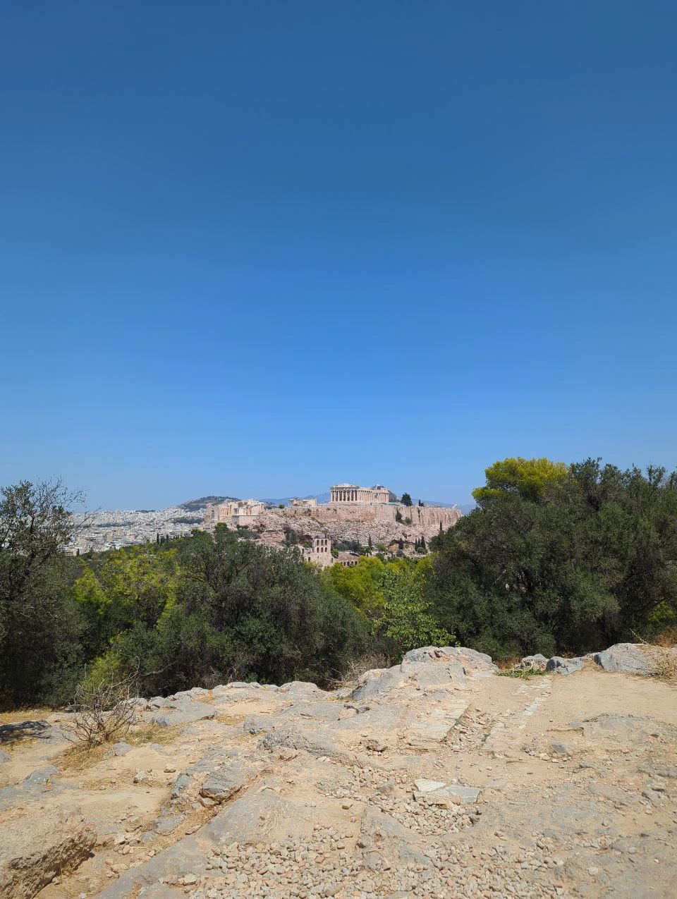 Acropolis Viewpoints - Rocky Philopappos Path An uneven rocky path on Philopappos Hill leads through trees toward a distant view of the Acropolis.