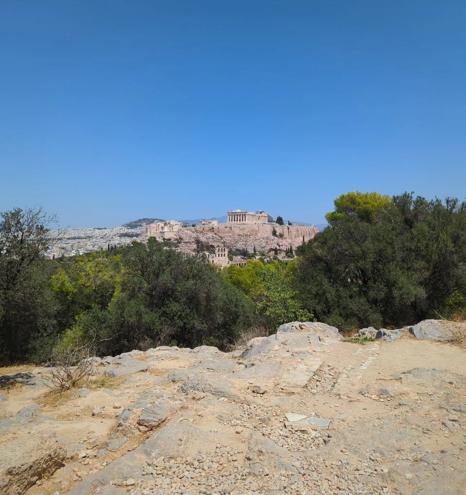 Athens Photoshoot - Rocky Philopappos Path An uneven rocky path on Philopappos Hill leads through trees towards the Acropolis, illustrating walking conditions for an Athens photoshoot.