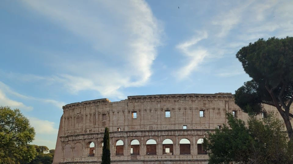 The Colosseum rises between umbrella pines and leafy trees beneath soft white clouds in Rome.
