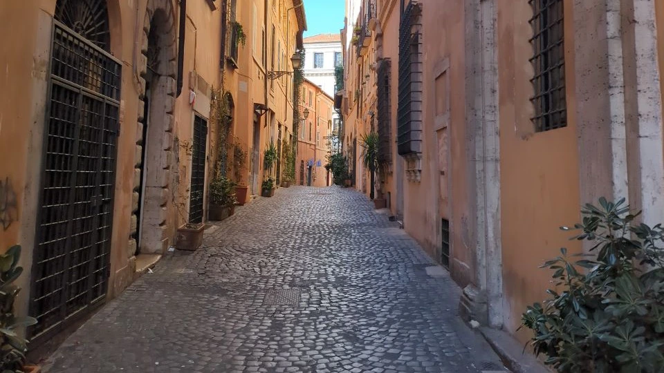 An empty cobbled lane runs between warm ochre façades and shuttered windows in Rome, creating a quiet start for a portrait route.