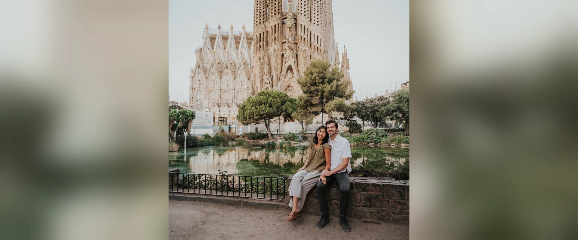 Two adults sit beside the pond at Sagrada Família with the basilica's towers rising behind them during a Barcelona photoshoot.