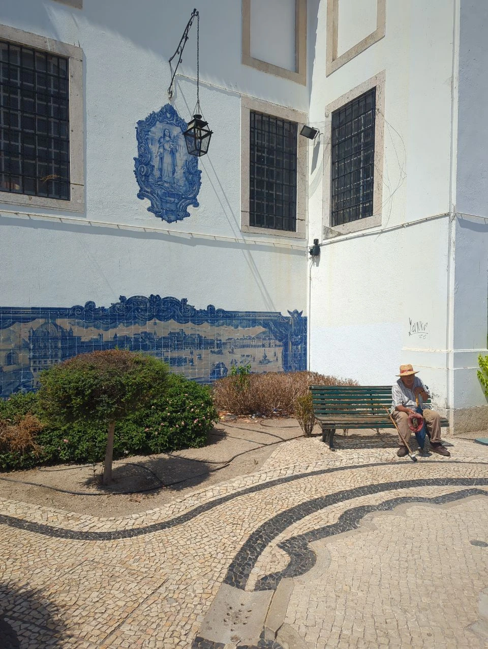 Lisbon Tram Photoshoot - Santa Luzia Terrace Blue-and-white azulejos, planted borders and patterned calçada surround a green bench with a seated visitor at Santa Luzia.
