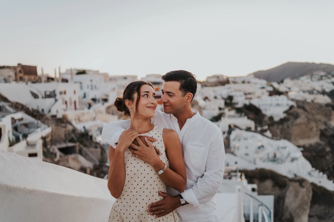 Santorini couple photoshoot: a smiling portrait among Oia’s white houses Man kissing his partner’s temple as she smiles with Oia’s white houses behind them during their Santorini couple photoshoot.