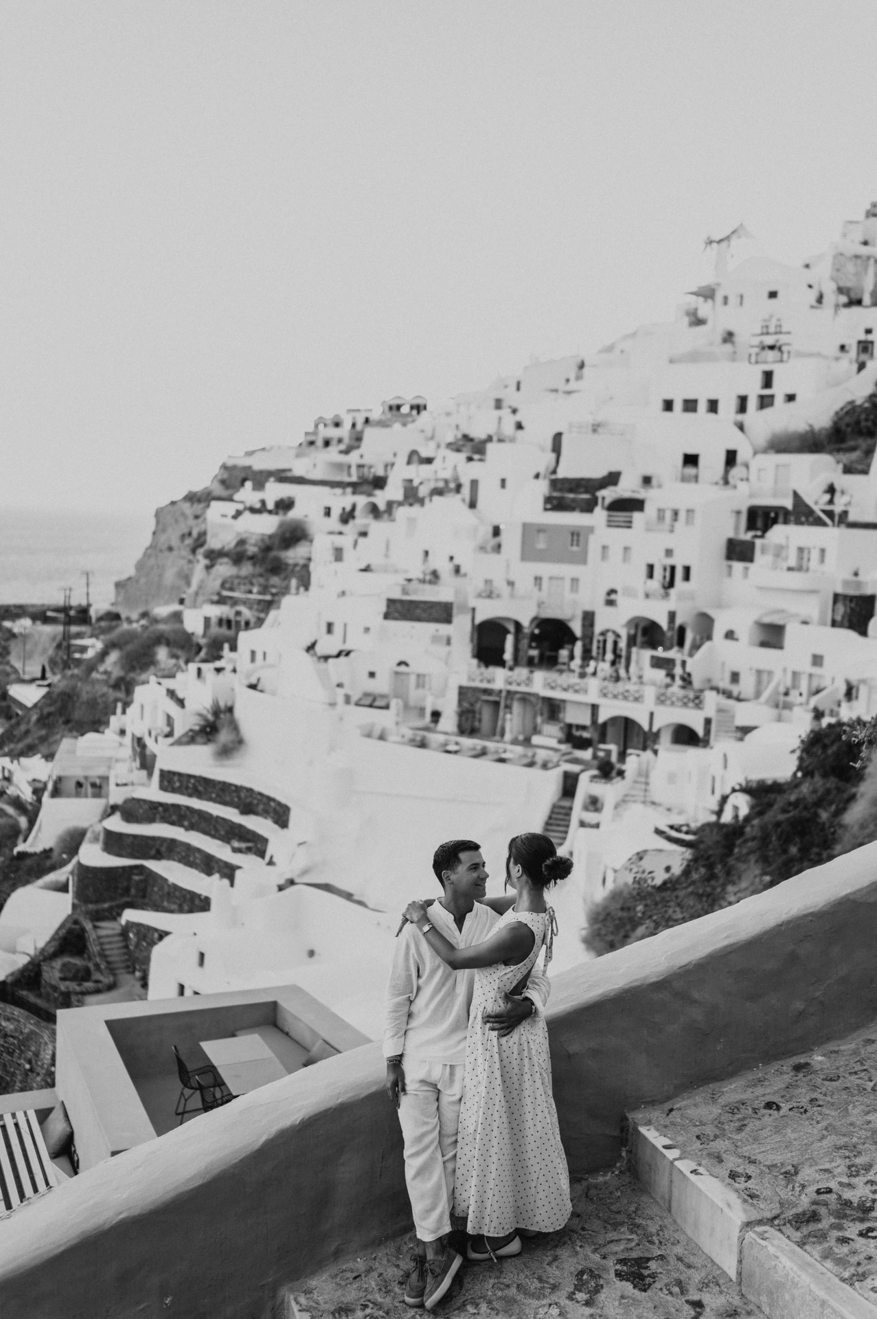 Black-and-white portrait of a couple embracing beside a white wall beneath Oia’s hillside houses during their Santorini couple photoshoot.