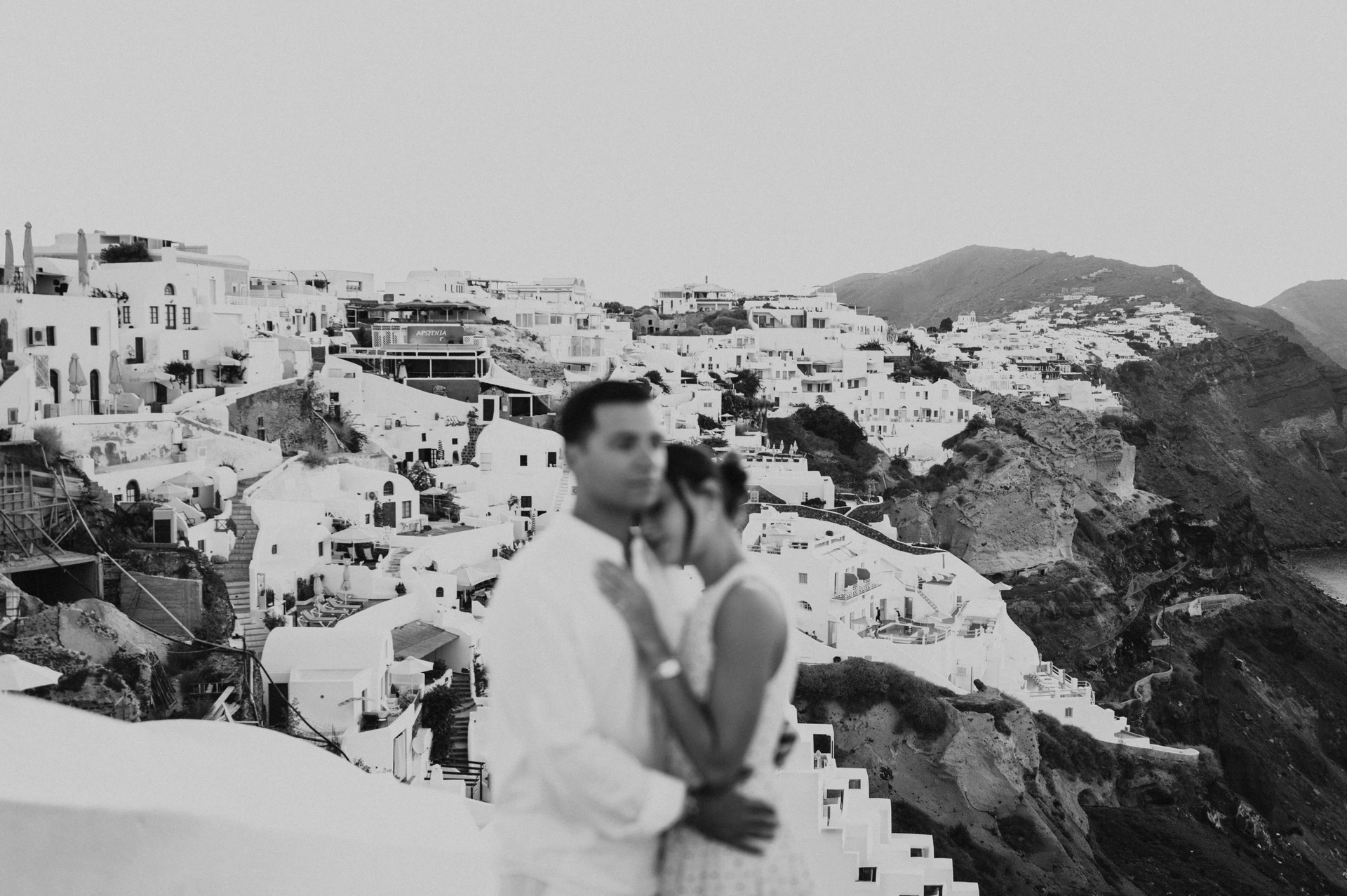 Black-and-white portrait of a couple embracing with white hillside buildings in the background during their Santorini couple photoshoot.