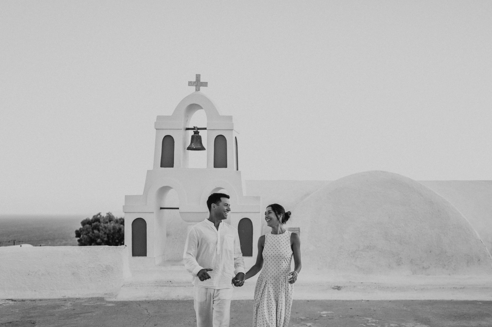 Black-and-white portrait of a couple standing beside a white bell tower during their Santorini couple photoshoot.
