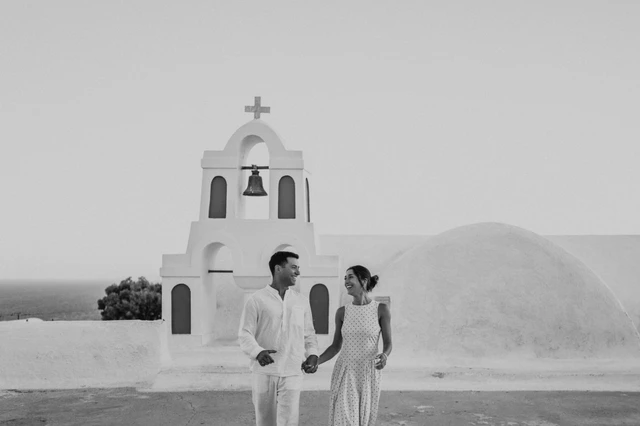 Black-and-white portrait of a couple standing beside a white bell tower during their Santorini couple photoshoot.