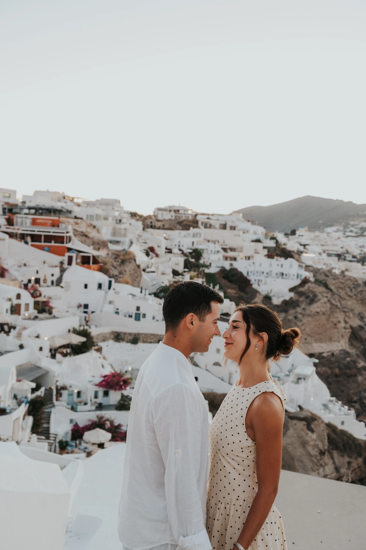 Couple standing close together beneath a broad sky above the sea and white hillside houses during their Santorini couple photoshoot.