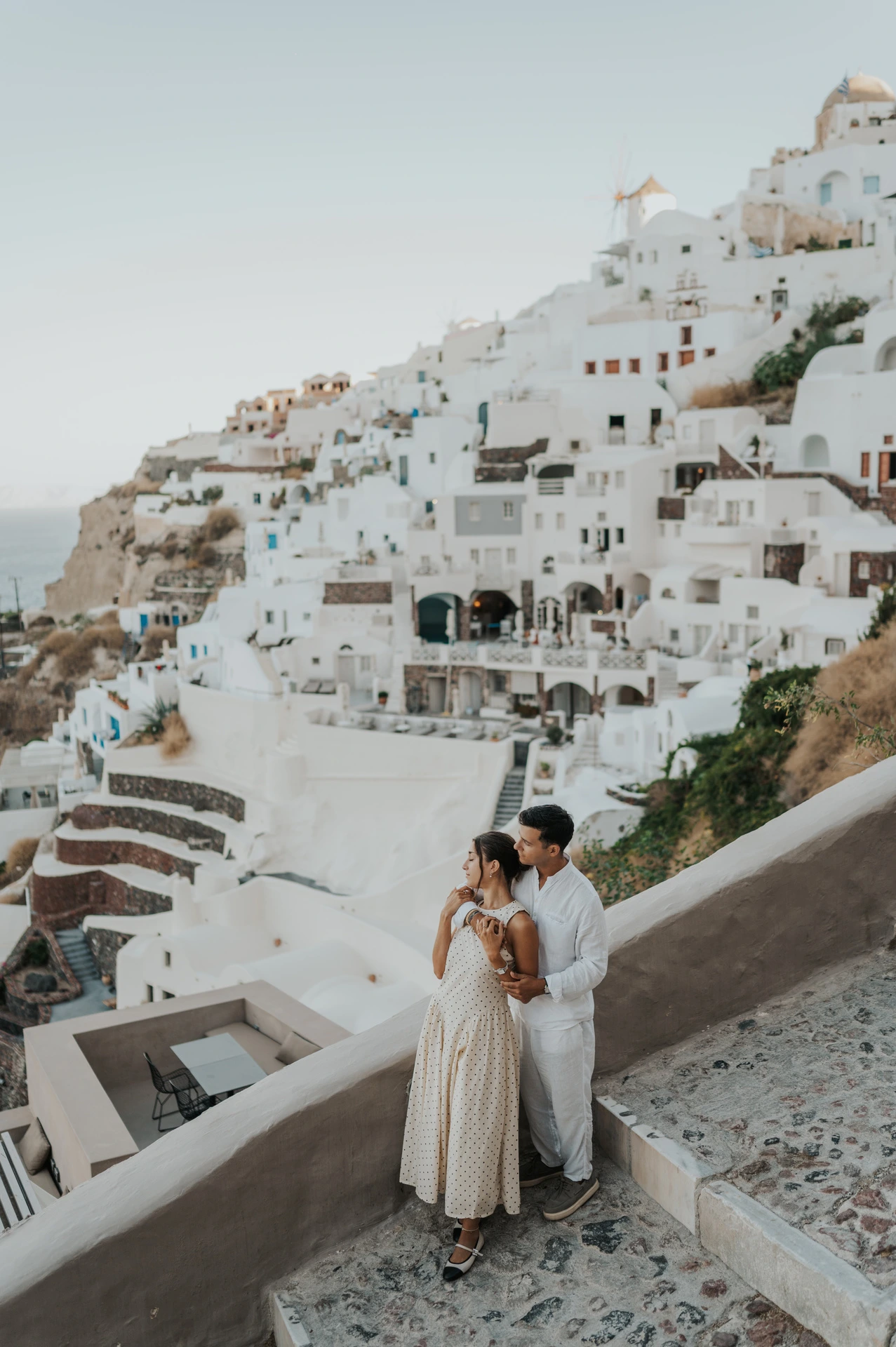 Couple standing together beside a sloping white wall with Oia’s hillside buildings behind them during their Santorini couple photoshoot.