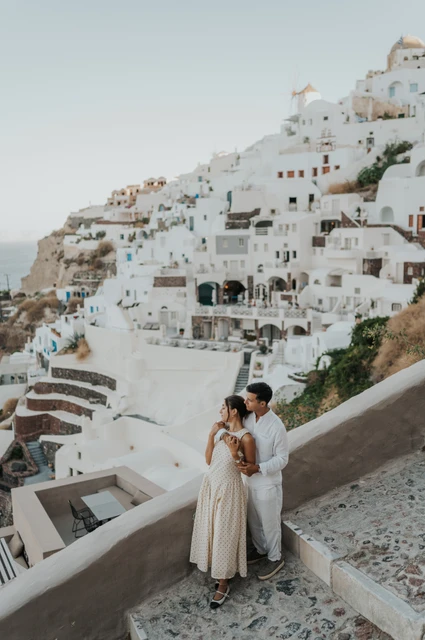 Santorini couple photoshoot: couple beneath Oia’s hillside buildings Couple standing together beside a sloping white wall with Oia’s hillside buildings behind them during their Santorini couple photoshoot.