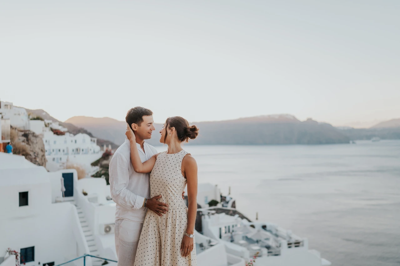 Santorini couple photoshoot: couple beside white walls and caldera cliffs Couple standing close beside white buildings with the sea and caldera cliffs behind them during their Santorini couple photoshoot.