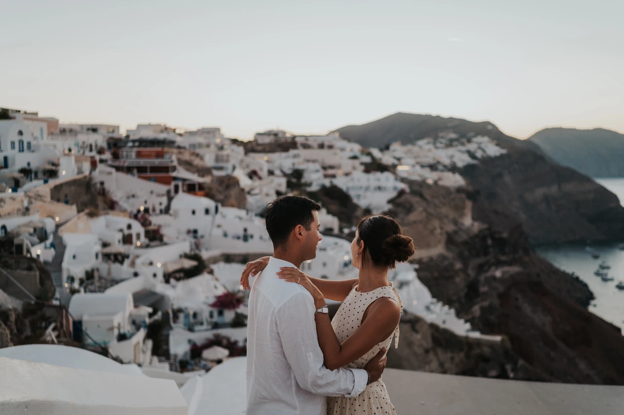 Santorini couple photoshoot: couple portrait above the caldera Couple facing each other beside a white wall with the caldera and hillside village behind them during their Santorini couple photoshoot.