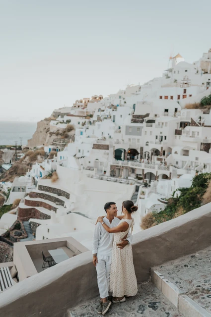 Santorini couple photoshoot: embrace beside Oia’s white hillside Couple embracing beside a sloping white wall below Oia’s white hillside houses during their Santorini couple photoshoot.