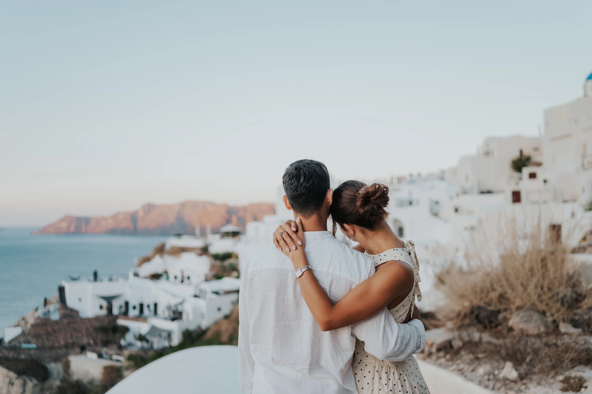 Couple with arms around each other looking towards the sea and distant caldera cliffs during their Santorini couple photoshoot.