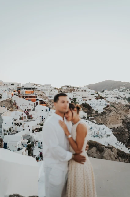 Santorini couple photoshoot: embrace with the hillside village behind Couple embracing and looking towards the camera with a white village across the hillside during their Santorini couple photoshoot.