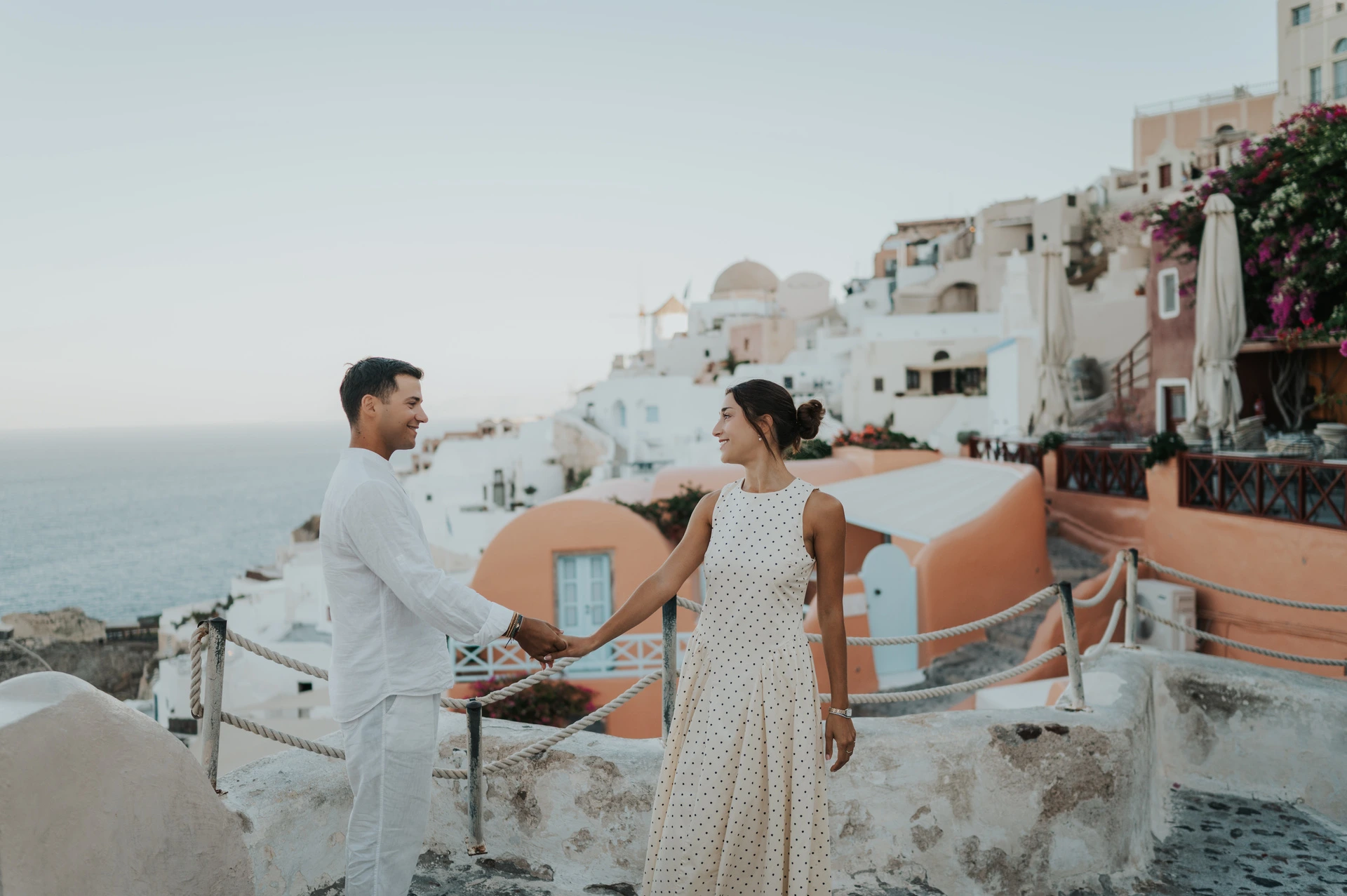 Couple holding hands on a cobbled terrace above peach-coloured buildings and the sea during their Santorini couple photoshoot.