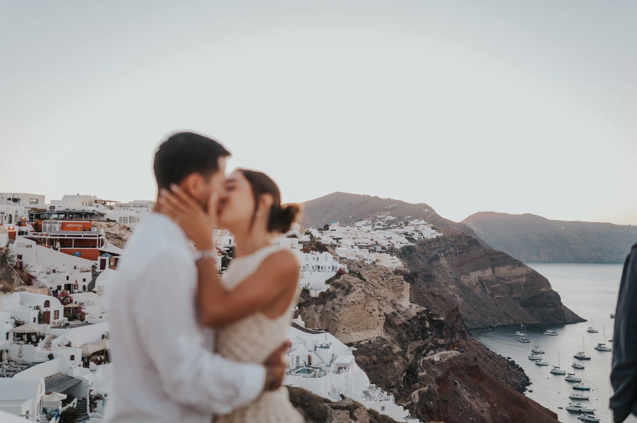 Santorini couple photoshoot: kiss beside a white caldera wall Wide portrait of a couple kissing beside a white wall above the caldera during their Santorini couple photoshoot.