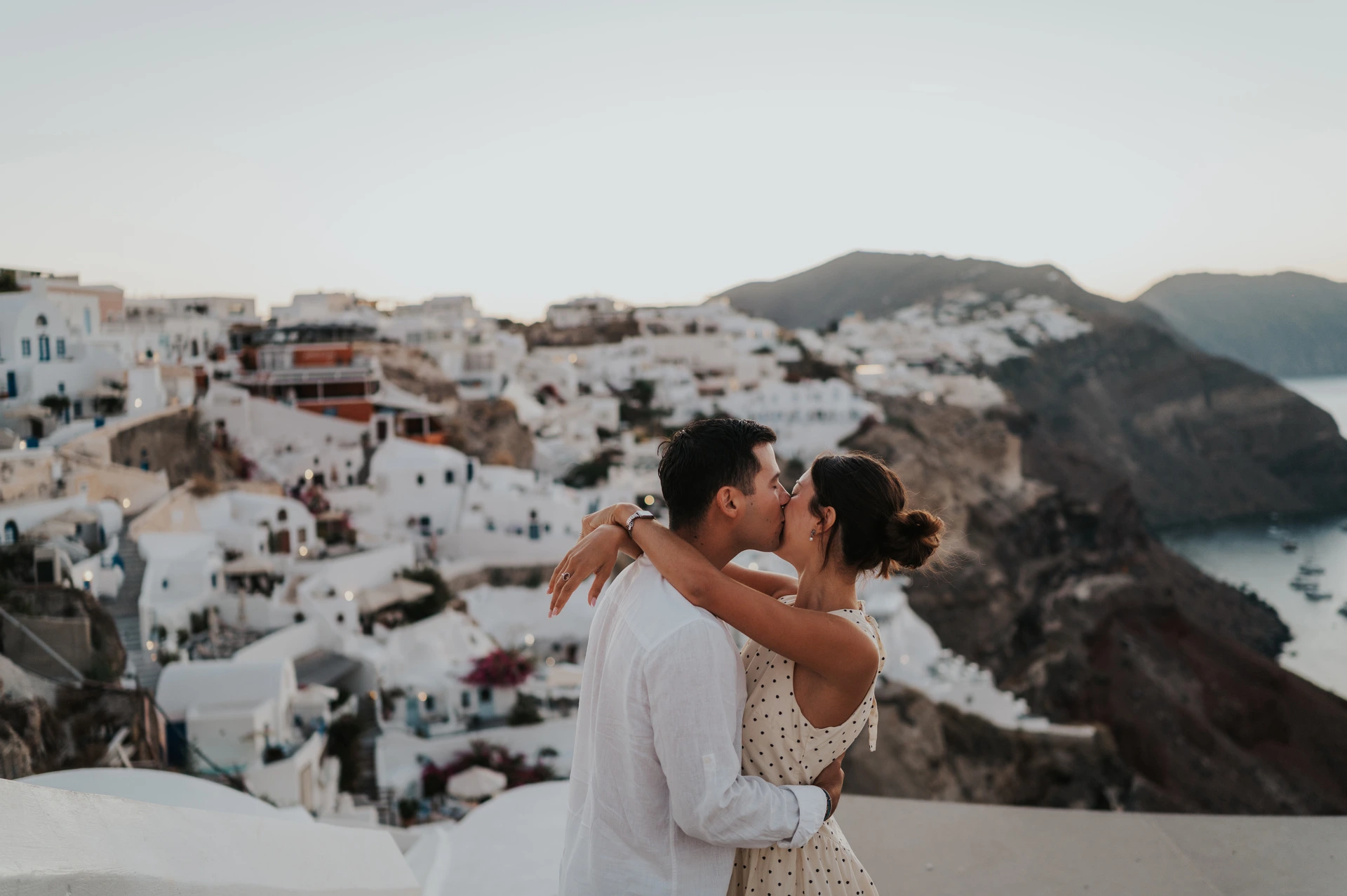 Couple kissing beside a white wall above the sea and hillside village during their Santorini couple photoshoot.