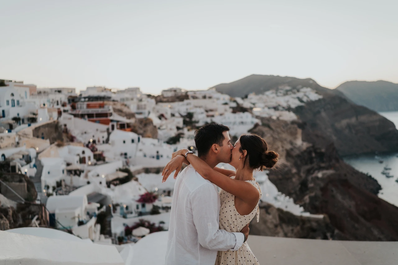 Santorini couple photoshoot: kiss overlooking the caldera Couple kissing beside a white wall above the sea and hillside village during their Santorini couple photoshoot.