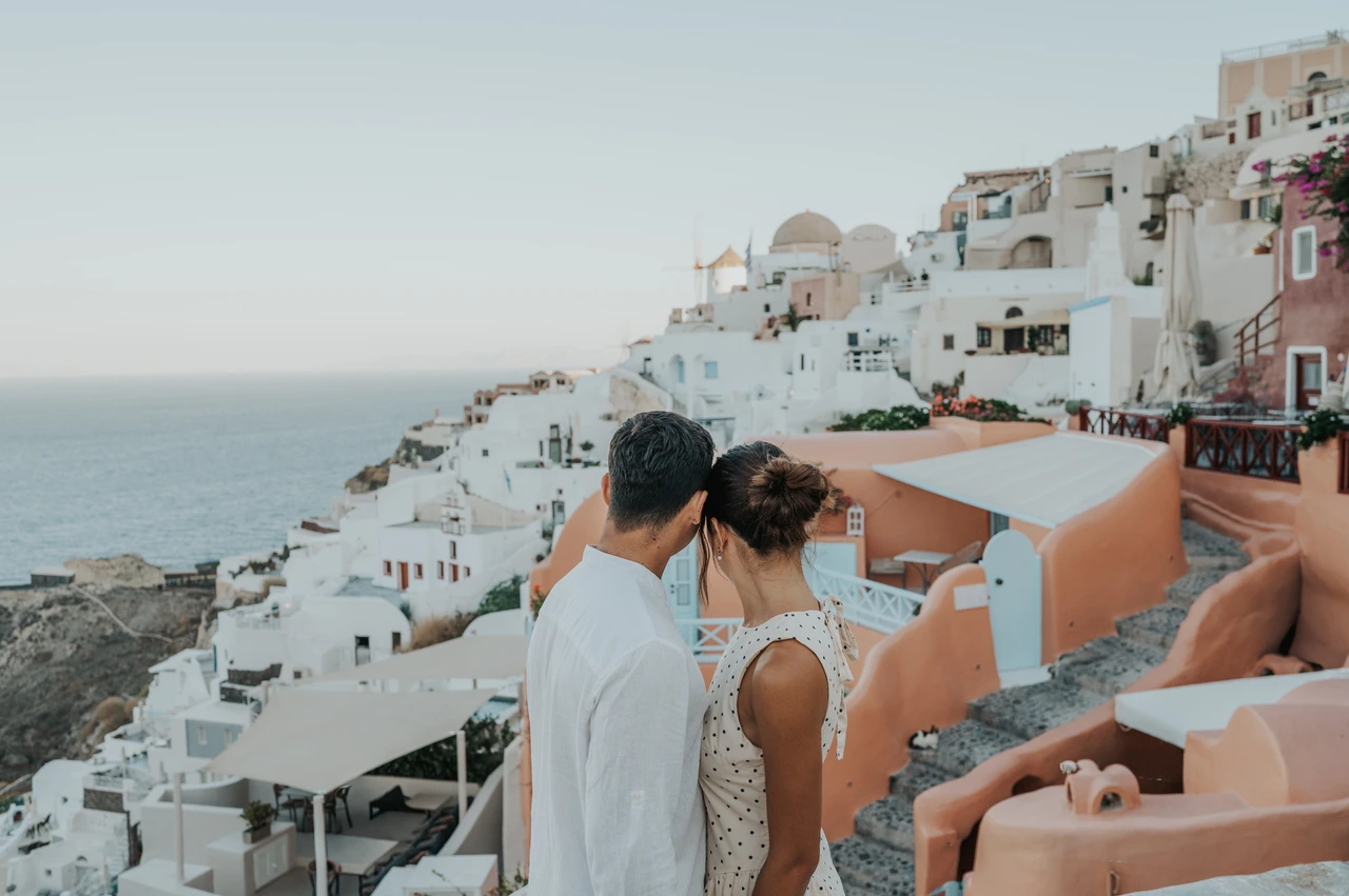 Santorini couple photoshoot: looking across Oia’s peach-coloured houses Couple seen from behind beside a curved white wall overlooking peach-coloured houses during their Santorini couple photoshoot.