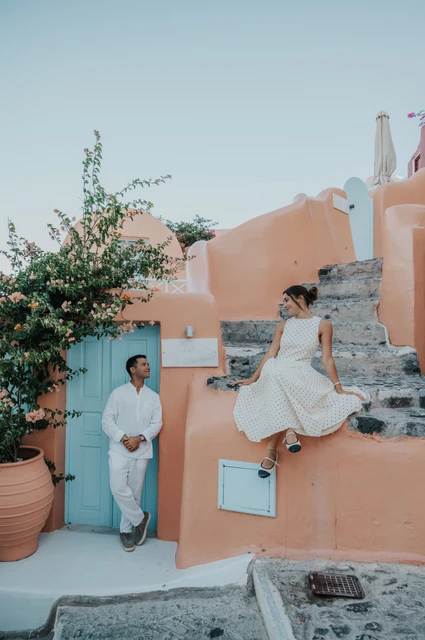 Woman lifting her skirt beside a peach-coloured wall while her partner stands by a pale blue door during their Santorini couple photoshoot.