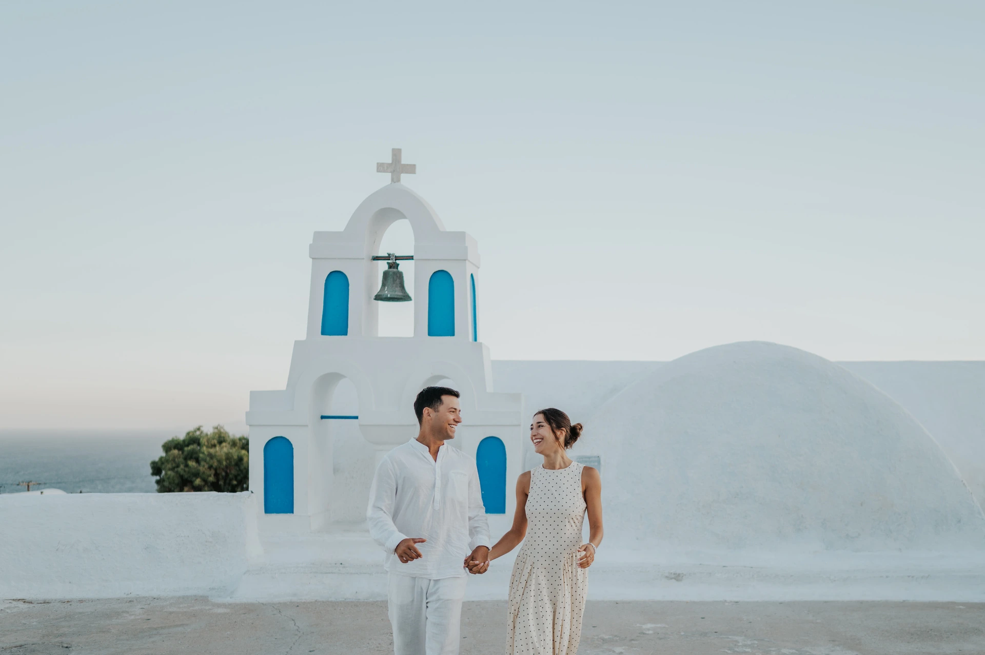 Couple standing close together beside a white bell tower with blue openings during their Santorini couple photoshoot.
