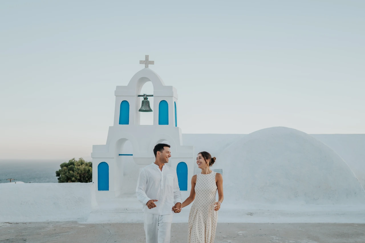 Santorini couple photoshoot: portrait beside a white and blue bell tower Couple standing close together beside a white bell tower with blue openings during their Santorini couple photoshoot.