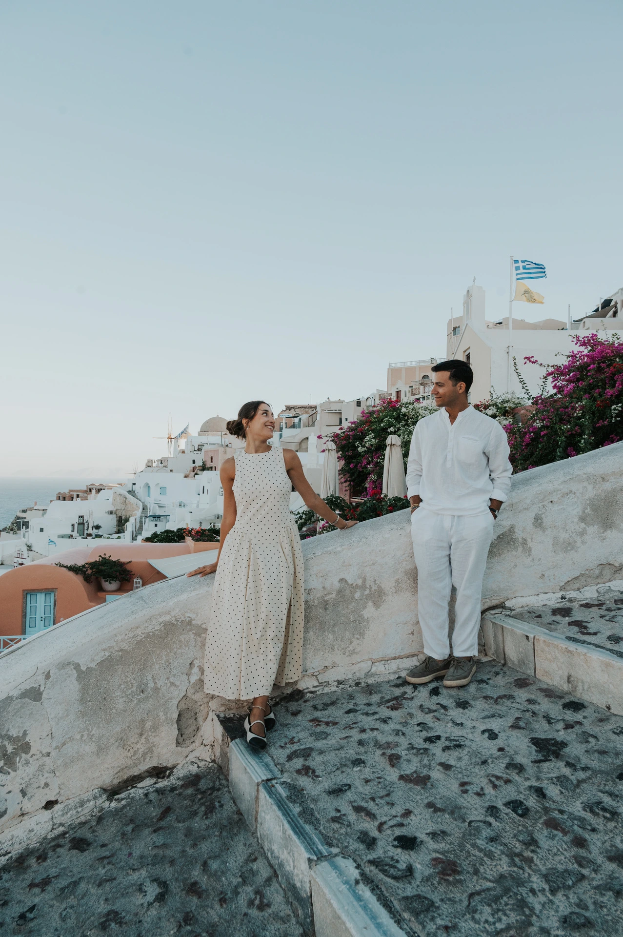 Full-length portrait of a couple holding hands beside white walls and peach-coloured buildings during their Santorini couple photoshoot.