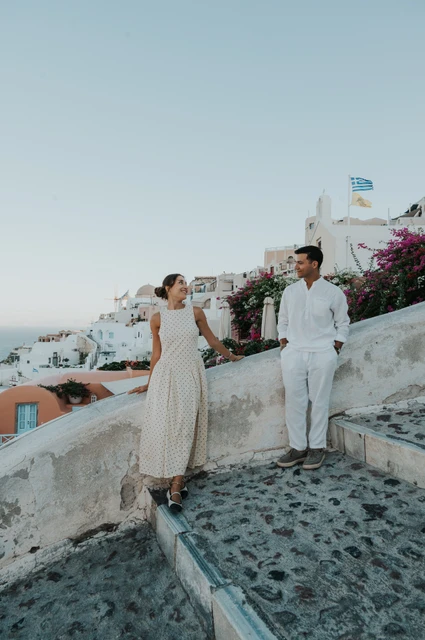Santorini couple photoshoot: portrait on Oia’s cobbled terrace Full-length portrait of a couple holding hands beside white walls and peach-coloured buildings during their Santorini couple photoshoot.