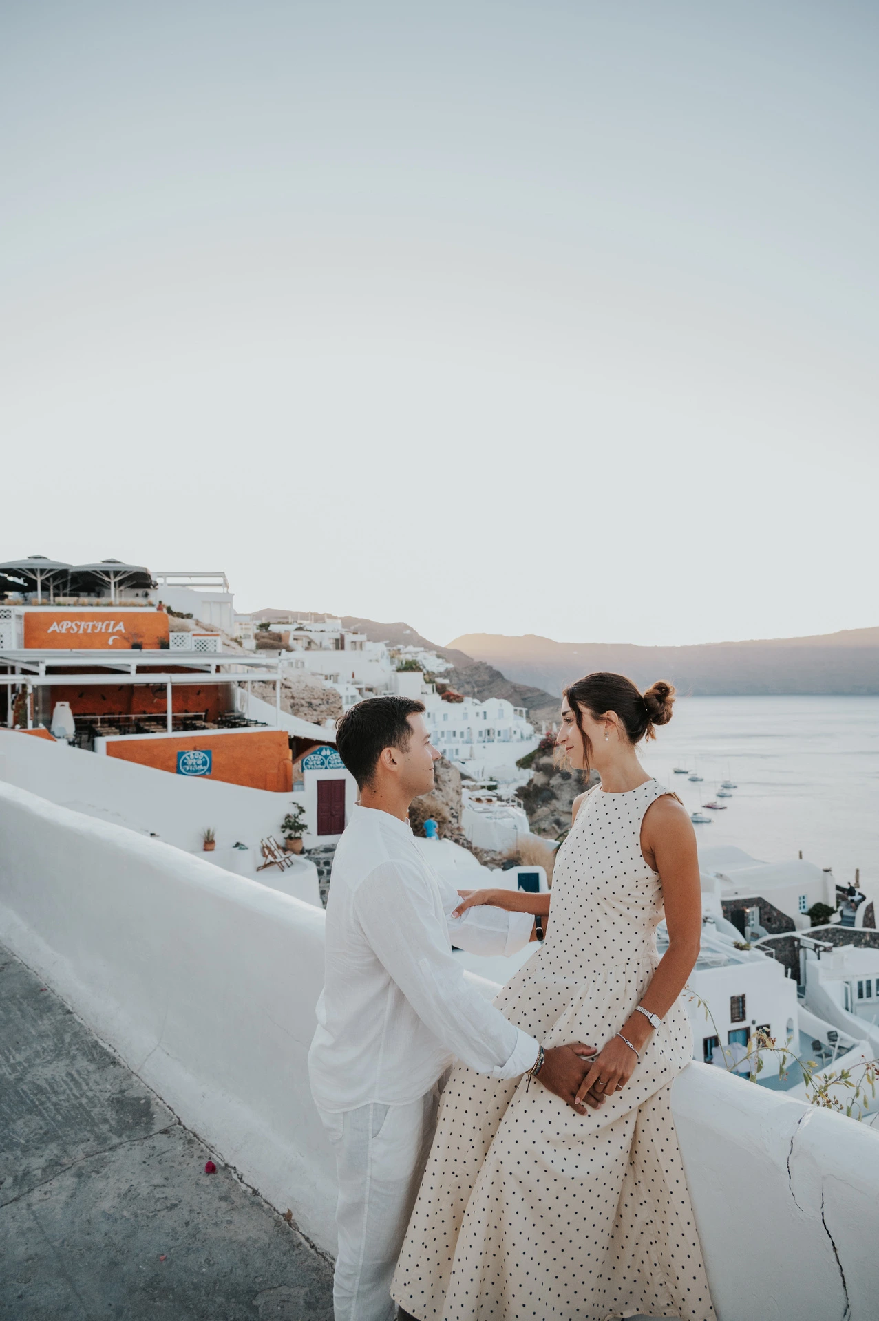Couple sitting together on a white wall with village buildings and the sea behind them during their Santorini couple photoshoot.