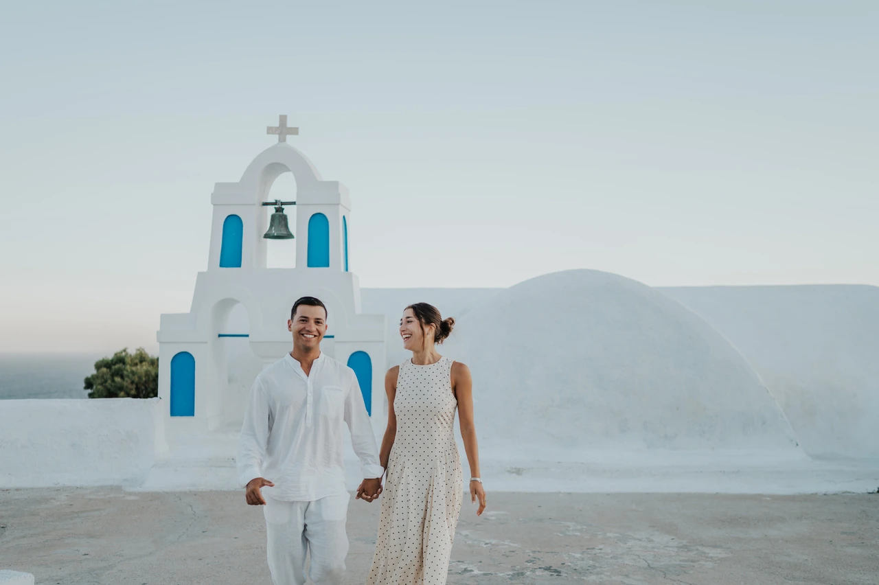 Santorini couple photoshoot: smiles beside the white bell tower Couple smiling together beside a white bell tower with blue openings during their Santorini couple photoshoot.
