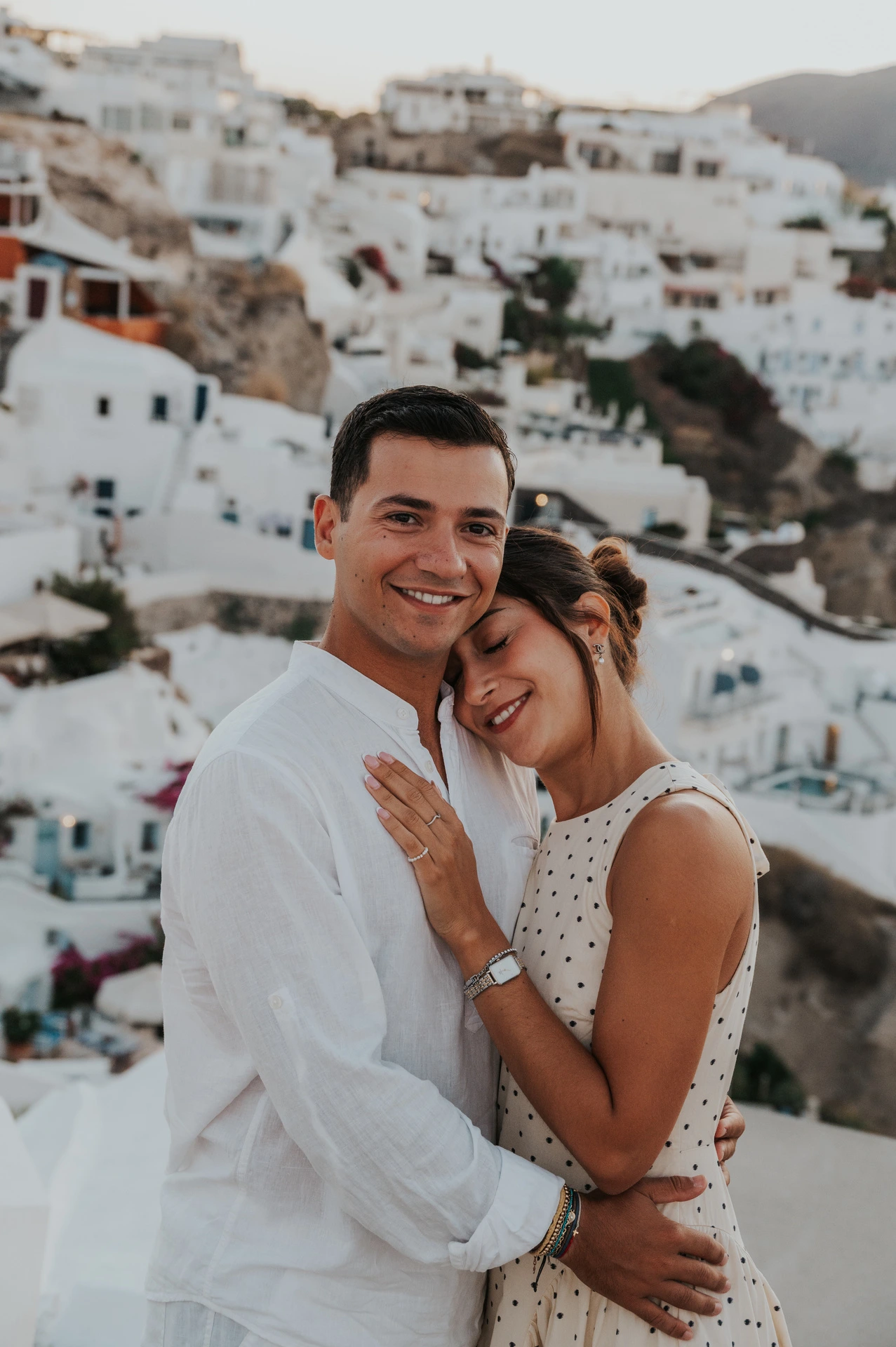 Close portrait of a smiling couple embracing above white hillside buildings during their Santorini couple photoshoot.