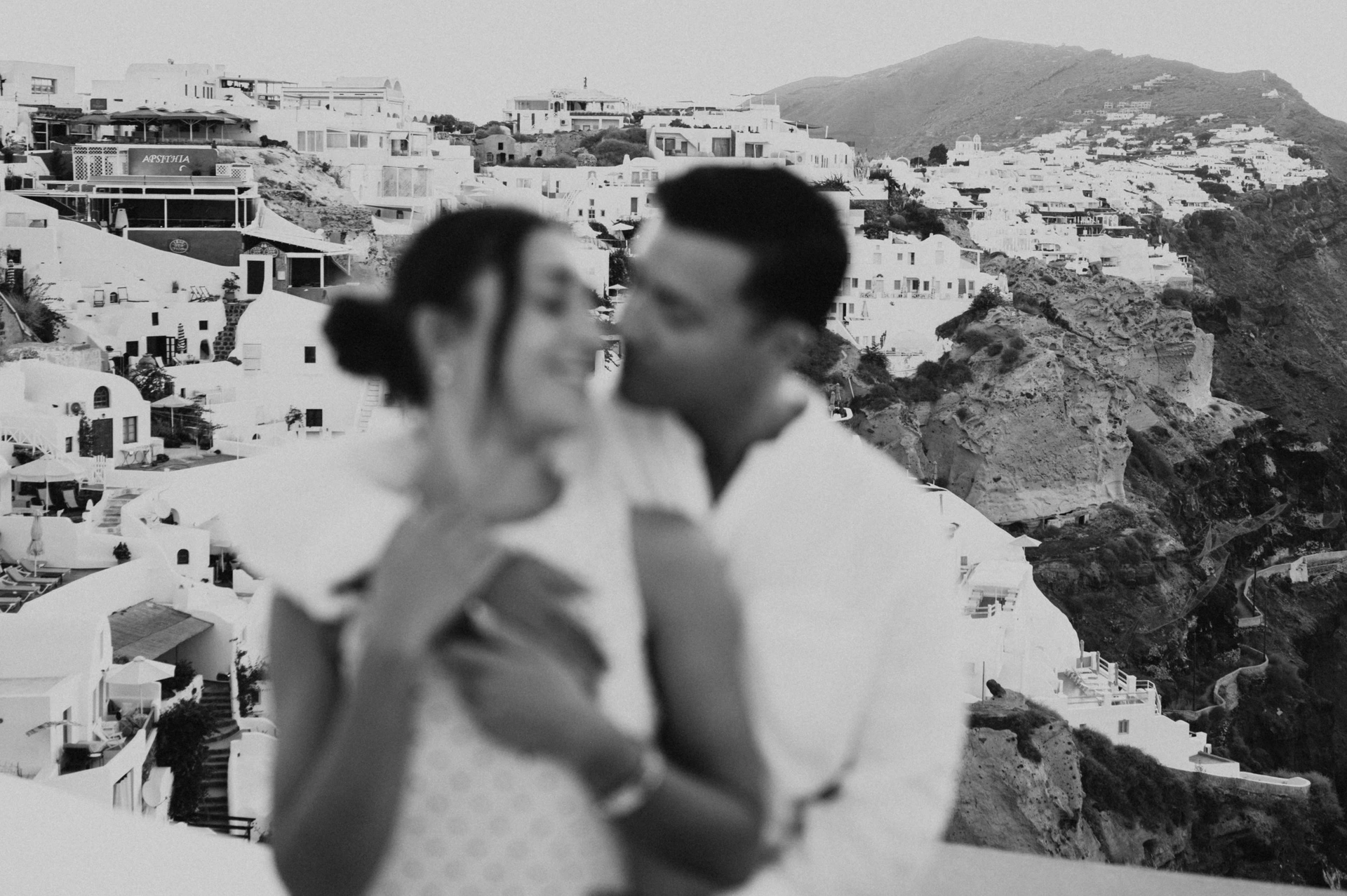 Soft-focus black-and-white portrait of a couple smiling together above the white village during their Santorini couple photoshoot.