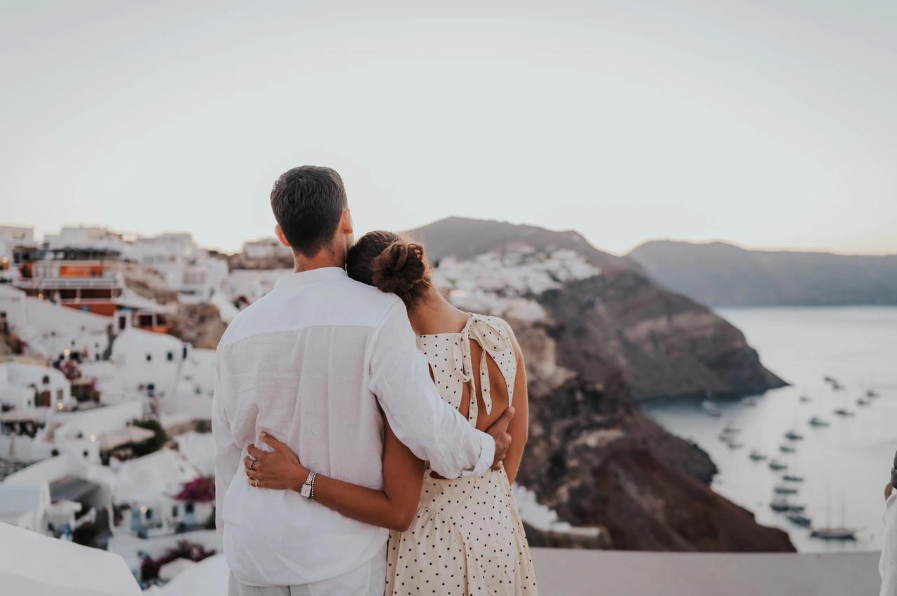 Santorini couple photoshoot: taking in Oia’s caldera view together Couple seen from behind with arms around each other above Oia’s white buildings and sea during their Santorini couple photoshoot.