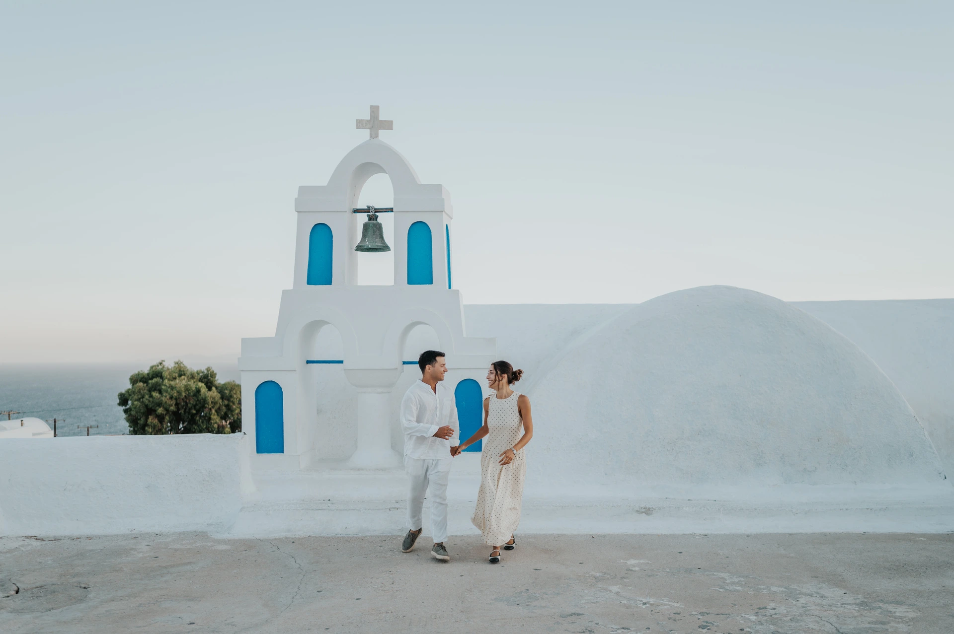 Couple walking hand in hand across a white terrace beside a bell tower with blue openings during their Santorini couple photoshoot.