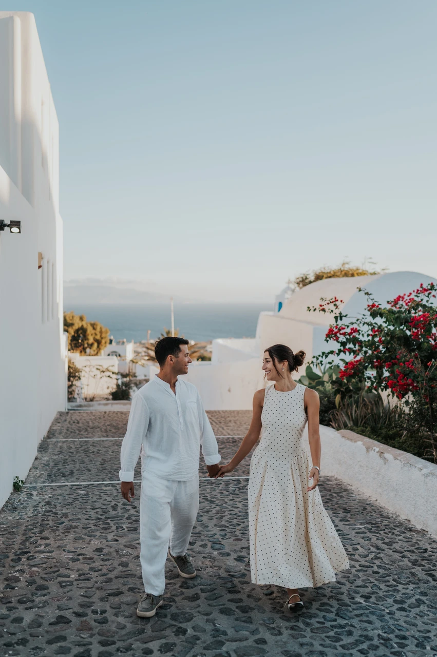 Santorini couple photoshoot: walking through a white village lane Couple walking hand in hand along a cobbled lane between white buildings and flowering plants during their Santorini couple photoshoot.