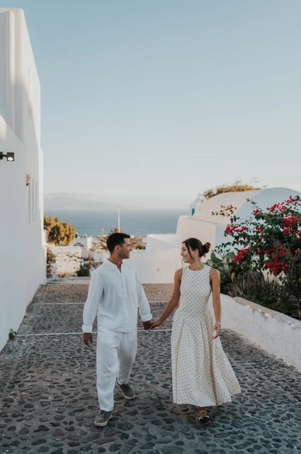 Santorini couple photoshoot: walking through a white village lane Couple walking hand in hand along a cobbled lane between white buildings and flowering plants during their Santorini couple photoshoot.